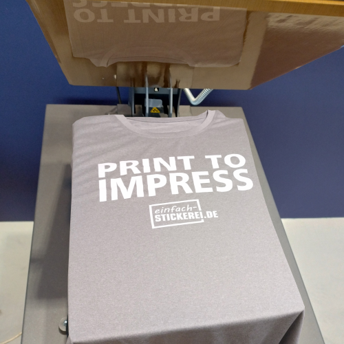 Graues T-Shirt in einer Transferpresse mit hochwertigem Textildruck. Der weiße Schriftzug 'PRINT TO IMPRESS' und das Logo 'einfach-STICKEREI.DE' werden mittels Thermotransfer-Druck aufgebracht. Professionelle Textilveredelung durch Druck und Stickerei.