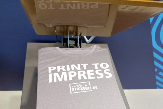 Graues T-Shirt in einer Transferpresse mit hochwertigem Textildruck. Der weiße Schriftzug 'PRINT TO IMPRESS' und das Logo 'einfach-STICKEREI.DE' werden mittels Thermotransfer-Druck aufgebracht. Professionelle Textilveredelung durch Druck und Stickerei.